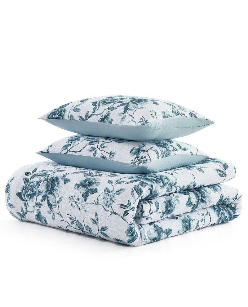 A folded bedding set with two matching pillows, featuring a white background with a dusty teal floral toile pattern and solid teal piping along the edges, set against a plain white background.