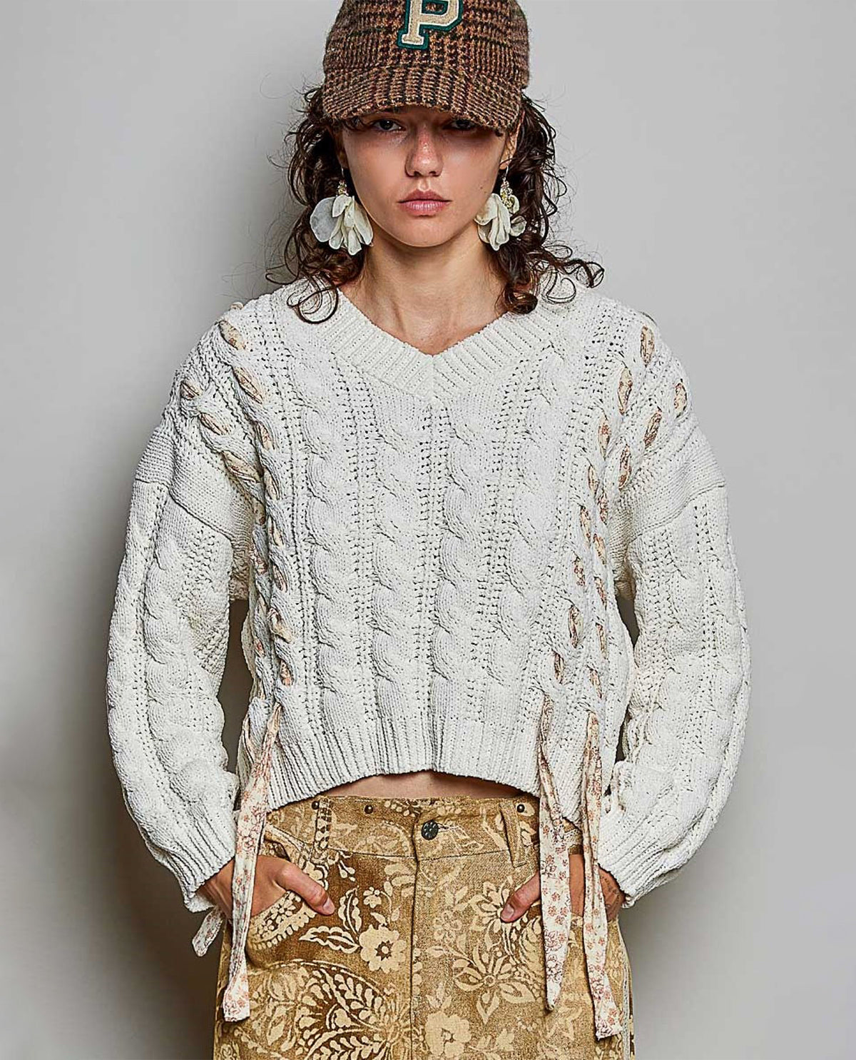 A woman wearing a cream cable-knit cropped sweater with side tie details, paired with a brown patterned cap and a beige floral print skirt, standing against a neutral gray background.