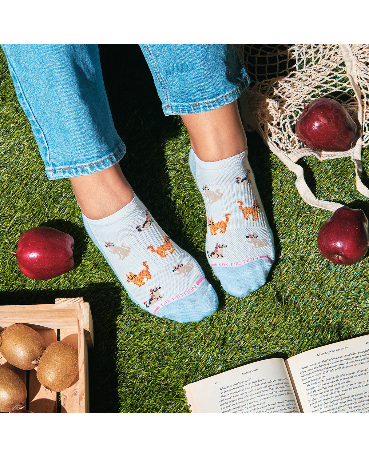 Person wearing light blue socks with animal patterns on grass, surrounded by apples and a book.