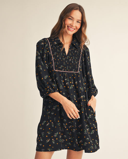 A woman modeling a black long-sleeve button-up dress with a small orange and green floral print and contrasting pink piping along the neckline, set against a plain beige studio background.