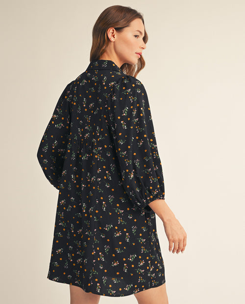 A woman wearing a black long-sleeve shirt dress with a small orange and white floral print, shown from a three-quarter back view against a light beige studio background.